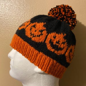 May include: A black knit beanie featuring a repeating orange jack-o'-lantern pattern around the base. The hat has a ribbed orange brim and is topped with a large, multicolored orange and black pom-pom, displayed on a white mannequin head.