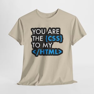 May include: A beige t-shirt featuring the text "YOU ARE THE {CSS} TO MY </HTML>". The text is in black and blue, with the CSS and HTML elements highlighted in blue. The shirt is made of a soft material.
