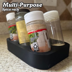 May include: A black, multi-purpose spice rack holding four spice jars. The jars are clear with white lids and contain various spices. The text "Multi-Purpose Spice rack" is at the top of the image.