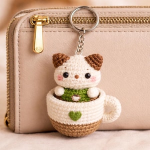 May include: A crocheted cat keyring in a coffee cup design. The cat is white and brown, with pink cheeks, and wears a green top. The cup is white and brown, with a green heart. The keyring is attached to a silver ring. The background is a beige purse.