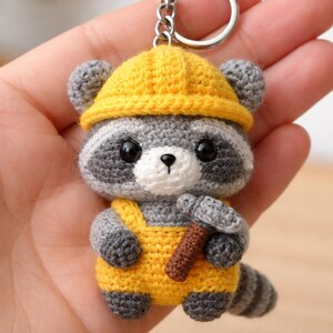 May include: A handmade crochet keychain featuring a raccoon wearing a yellow construction hat and overalls. The raccoon has gray and white fur, black eyes, and a small brown hammer. The keychain is attached to a silver ring.