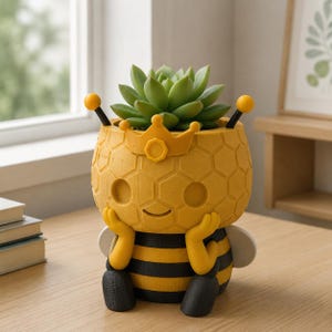 Può includere: Un vaso a forma di ape giallo e nero con una texture a nido d'ape e una piccola corona sul davanti. Il vaso presenta due antenne con punte gialle, due braccia appoggiate contro il viso e una pianta succulenta verde nell'apertura superiore.