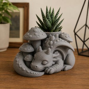 May include: A grey 3D-printed planter featuring a sleeping dragon curled around several mushrooms. The dragon has textured scales and closed eyes, with a central opening designed to hold a small green succulent plant with white striped leaves.