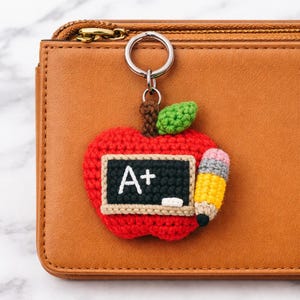 May include: A red crocheted apple keychain with a black chalkboard displaying "A+" and a pencil. The apple has a green leaf and a brown stem. The keychain is attached to a silver ring.