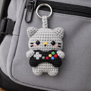 May include: Crocheted grey cat keyring holding a black video game controller with colourful buttons. The cat has pink cheeks, black eyes, and a yellow nose. The keyring is attached to a silver metal ring.