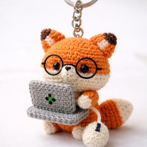 Puede incluir: Llavero de zorro de ganchillo con detalles en naranja, blanco y marrón. El zorro lleva gafas redondas negras y sostiene un portátil gris con un símbolo de cruz verde. Un ratón blanco está conectado al portátil.