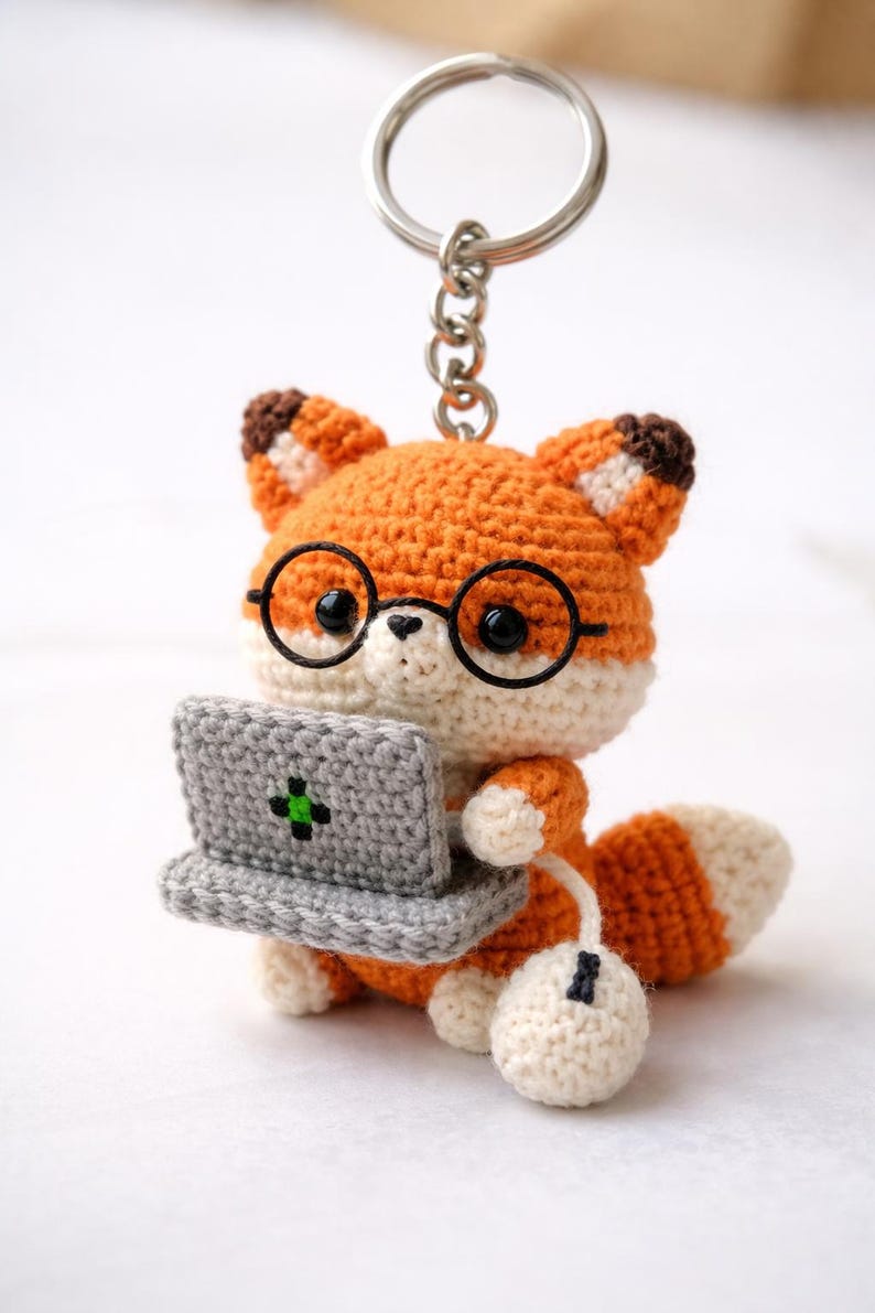 May include: Crocheted fox keychain with orange, white, and brown details. The fox wears round black glasses and holds a gray laptop with a green cross symbol. A white mouse is connected to the laptop. The keychain is attached to a silver ring.