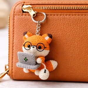 May include: A crocheted fox keychain in orange, white, and brown, wearing glasses and holding a gray laptop and a white mouse. The fox is attached to a silver keyring, set against a brown leather wallet.