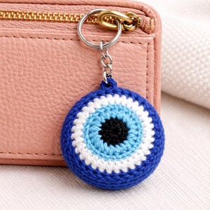 May include: A blue, white, and black crochet evil eye keychain. The keychain is round with a blue outer ring, white and light blue inner rings, and a black center. It hangs from a silver keyring attached to a gold zipper.