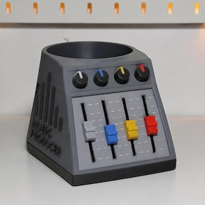 May include: A dark gray desk organizer shaped like a music mixing console with a circular top opening. The front features four black knobs with white, blue, yellow, and red indicators, and four vertical sliders in gray, blue, yellow, and red.