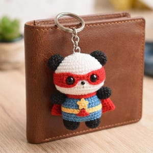 Könnte beinhalten: Ein handgefertigter Panda-Schlüsselanhänger im Superhelden-Kostüm. Der Panda hat einen weißen Kopf mit schwarzen Ohren, eine rote Maske und ein blau-gelbes Outfit. Der Schlüsselanhänger ist an einem silbernen Metallring befestigt.