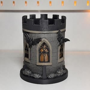 May include: A cylindrical castle tower pen holder featuring a dark gray stone brick texture, a crenelated top, and three arched windows with gold-colored panes. Two black bat figures are attached to the exterior walls, and the base is circular.