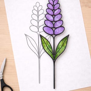 Puede incluir: Una flor de lavanda de vidriera con pétalos morados y hojas verdes, colocada sobre un papel blanco con un contorno dibujado. Unas tijeras negras descansan sobre una superficie de madera.