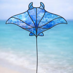 May include: A stained glass manta ray sculpture with a blue and white marbled design. The sculpture has a black outline and a long, thin black stem. The background is a blurred image of a beach and ocean.
