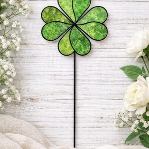 Puede incluir: Una estaca de jardín con forma de trébol de cuatro hojas con un tallo negro y un diseño de trébol verde texturizado. El trébol está rodeado de flores blancas y se encuentra sobre un fondo de madera blanca.