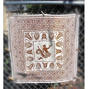 以下が含まれることがあります： 屋外に展示されている、白と茶色の模様のテキスタイル。スカーフまたは壁掛けの可能性があります。デザインは、繰り返しの幾何学的なボーダーと、三叉の槍を持った人物の画像が特徴です。テキスタイルは、洗濯ばさみで金網のフェンスに固定されています。