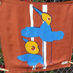 May include: A rust-colored textile with a whimsical bird design. Two blue birds with yellow heads and white accents are featured against a backdrop of white vertical stripes. The textile is held by wooden clothespins.