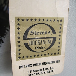 May include: A vintage fabric tag with the text "Stevens HOCKANUM Fabrics" in a black and gold design. The tag reads "FINE FABRICS MADE IN AMERICA SINCE 1813" and includes the address of J.P. Stevens & Co. in New York.