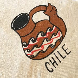 Puede incluir: Una tela beige con una ilustración marrón de una jarra tradicional con asa y una figura animal estilizada. La jarra tiene patrones decorativos negros, rojos y blancos. La palabra "CHILE" está impresa en negro debajo.