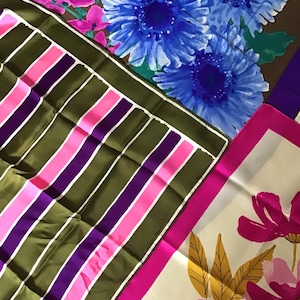 May include: A collection of three square scarves featuring distinct patterns. One scarf displays olive green, purple, and pink vertical stripes. Another scarf shows blue flowers on a brown background, and the third features pink flowers on white.