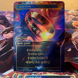 May include: A collectible trading card featuring a golden ring with intricate inscriptions. The card has a holographic design with a blue and purple background. The text "1 of 1 Ring" is visible, along with the artist's name and the year 2023.