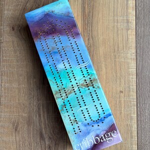 Op de afbeelding: Een rechthoekig cribbagebord met een abstract ontwerp in blauw, turquoise, paars en goud. Het bord heeft veel kleine gaatjes voor het plaatsen van pinnen. Het woord "cribbage" staat in witte letters.