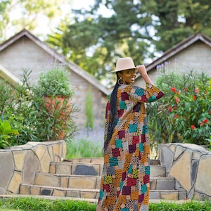 May include: A woman wearing a long, patterned dress with a beige hat. The dress features a patchwork design in shades of red, yellow, green, and blue. She is standing on a grassy area with stone steps and buildings in the background.