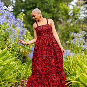 Könnte beinhalten: Ein rotes und schwarzes gemustertes Maxikleid mit quadratischem Ausschnitt und Spaghettiträgern. Das Kleid hat einen gerüschten Saum und wird im Freien zwischen blauen Blumen und grünem Laub getragen.