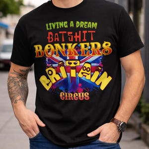 Puede incluir: Camiseta negra con un diseño gráfico colorido. El diseño incluye el texto "LIVING A DREAM BATSHIT BONKERS BRITAIN CIRCUS" en varias fuentes y colores, con un fondo de la bandera británica.