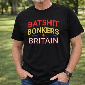 Puede incluir: Camiseta negra con las palabras "BATSHIT BONKERS BRITAIN" en texto rojo, amarillo y blanco. La palabra "BONKERS" tiene un gráfico de estrella. La camiseta la lleva una persona con vaqueros azules y un reloj.