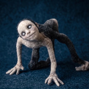 May include: A needle-felted creature figurine with a pale, textured face, bald head, and solid white eyes. The body is a mix of light beige and dark charcoal gray fibers, featuring long, thin limbs and a hunched, quadrupedal posture.
