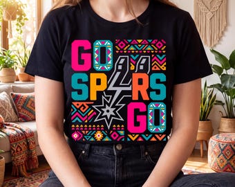 May include: Black t-shirt with the words "GO SPURS GO" in bright pink, orange, and teal letters. The design includes a stylized lightning bolt and a star, with colorful geometric patterns. The shirt is a casual wear item.