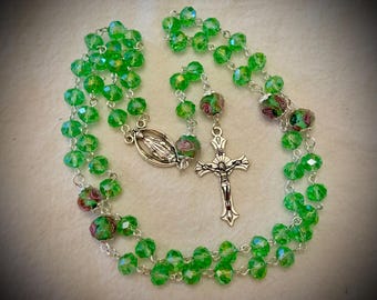 May include: A rosary featuring translucent bright green faceted beads linked by silver-toned metal chain. The piece includes a silver-toned crucifix and a central oval medal depicting a figure, interspersed with floral-patterned beads.