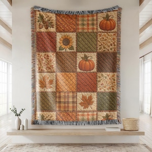 May include: A woven throw blanket featuring a patchwork design with autumn-themed squares. The pattern includes orange pumpkins, sunflowers, maple leaves, acorns, and plaid textures in shades of brown, green, and orange with fringed edges.