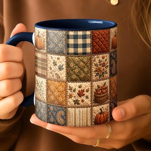 May include: A ceramic mug with a dark blue handle and interior, featuring a quilted patchwork pattern on the exterior. The design includes squares with pumpkins, blue and white gingham, floral motifs, autumn leaves, and striped fabric textures.