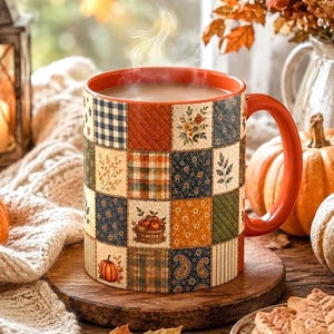 May include: A ceramic mug featuring a colorful patchwork pattern of autumn-themed squares, including pumpkins, flowers, and plaid designs. The mug has a solid orange interior and handle, and it is filled with a steaming hot beverage.