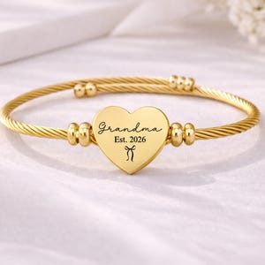 Pode incluir: Uma pulseira de fio dourado trançado com um pingente central em forma de coração. O coração tem gravado o texto "Grandma", "Est. 2026" e um pequeno ícone de laço. A pulseira inclui duas pequenas contas douradas em cada lado do coração.