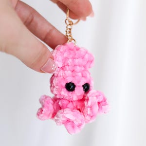 May include: A handmade pink crochet octopus keychain with black eyes and a gold-colored keyring. The plush toy is held up against a white background. The octopus has eight tentacles and a rounded head.