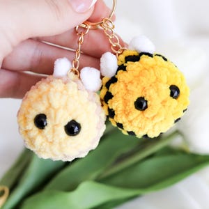 Puede incluir: Dos llaveros de abeja hechos a mano con ganchillo. Uno es amarillo con rayas negras, el otro es amarillo claro. Ambos tienen ojos negros y antenas blancas, unidos a llaveros dorados. Los llaveros son suaves y esponjosos.