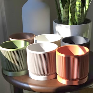 May include: A collection of cylindrical planters in various colors, including green, brown, white, and orange. Each planter has vertical ribbed detailing and sits on a small, matching base. A large white vase and a white planter with a snake plant are in the background.
