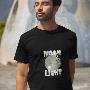 May include: Black t-shirt with a graphic of a full moon and the words "MOON LIGHT" in white. The shirt is worn by a person standing outdoors.