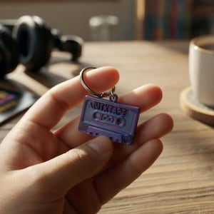 Peut inclure: Un porte-clés en forme de cassette audio violet et bleu avec le mot "AMIXTAPE". Le porte-clés est tenu dans une main. En arrière-plan, un casque noir, une tasse de café et un disque vinyle.