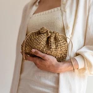 May include: A woven straw drawstring bag in a natural tan color. The bag has a rounded shape and a drawstring closure. The bag is held by a person wearing a cream-colored outfit.