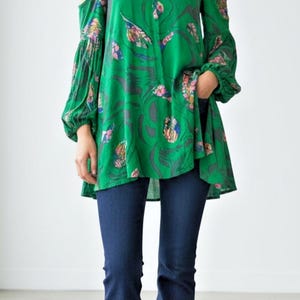 May include: A vibrant green cold-shoulder tunic featuring a high neckline, long billowy sleeves with gathered cuffs, and a multi-colored abstract floral pattern. The tunic is paired with dark blue denim flare jeans and simple black flat shoes.