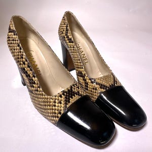 May include: A pair of Chanel pumps featuring a snakeskin-patterned body in shades of tan and dark brown, complemented by a solid black patent leather cap toe. The shoes have a block heel and the Chanel logo is printed in gold on the inner sole.