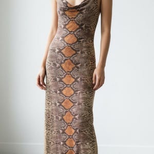 Pode incluir: Um vestido comprido sem mangas com estampado de pele de cobra em tons de castanho e bege, com um padrão de diamantes laranja ao centro. A peça apresenta um decote drapeado e alças finas, combinado com botas pretas.