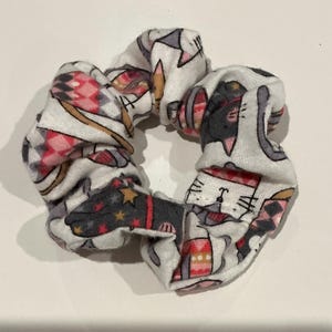 Könnte beinhalten: Ein weißes Haargummi mit einem sich wiederholenden Muster aus grauen Katzen, roten und rosa geometrischen Formen und goldenen Sternen. Dieses Haaraccessoire ist für einen Pferdeschwanz oder Dutt konzipiert.