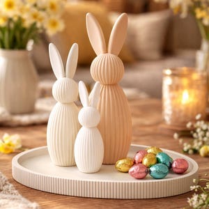 Op de afbeelding: Drie decoratieve paashaasbeeldjes in crème- en perziktinten staan op een wit dienblad met in folie verpakte chocolade-eieren. De konijnen hebben lange oren en geribbelde lichamen. De scène speelt zich af op een houten tafel met bloemen en een kaars.