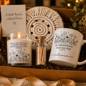 May include: A gift box containing a white mug with floral line art and the text be still and know that I am God, a lit candle with floral art and religious text, a small glass bottle of matches, a round macrame coaster, and a note card.
