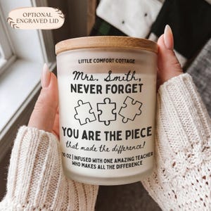 May include: A frosted glass candle jar with a bamboo lid featuring black text that reads: LITTLE COMFORT COTTAGE, Mrs. Smith, NEVER FORGET YOU ARE THE PIECE that made the difference! 10 OZ | INFUSED WITH ONE AMAZING TEACHER WHO MAKES ALL THE DIFFERENCE.